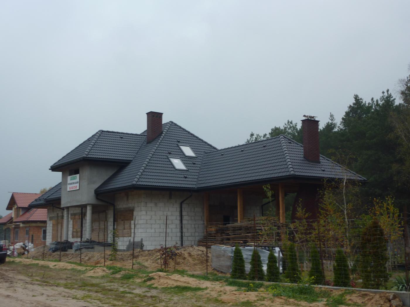 Dom jednorodzinny w trakcie budowy z nowym dachem z ciemnej dachówki, widoczne kominy, okna dachowe i częściowo wykończona elewacja z białego pustaka. Na elewacji wisi baner reklamowy.